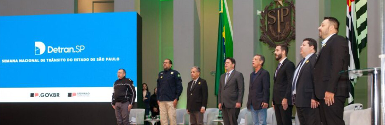A imagem mostra um evento com várias pessoas no palco. No centro do palco, há uma grande tela exibindo a marca do “Detran-SP” e, abaixo, as palavras, em maiúsculas: “SEMANA NACIONAL DE TRÂNSITO DO ESTADO DE SÃO PAULO”. Há logotipos no canto inferior da tela, possivelmente de patrocinadores ou parceiros do evento. A tela é cercada por três painéis verticais amarelos de cada lado e o fundo do palco é azul-escuro. Há várias bandeiras em mastros, incluindo a do estado de São Paulo, à direita. No palco, há dez indivíduos, cinco de cada lado da tela, em frente a um pódio com um orador. Eles estão de pé e parecem prestar atenção no orador.