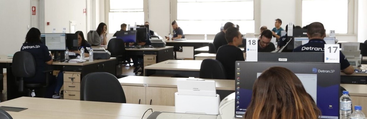 A imagem mostra um ambiente de escritório amplo. Em primeiro plano, à esquerda, um homem com camisa azul está sentado de perfil, trabalhando em um computador. Atrás dele, uma mulher também está sentada de frente para um computador. Várias mesas com computadores estão dispostas em filas ao longo do escritório, e várias pessoas estão trabalhando nelas. Os móveis são predominantemente pretos e beges. Cada estação de trabalho está numerada, e os monitores exibem o logotipo do Detran-SP. Há várias janelas ao fundo, permitindo a entrada de luz natural.