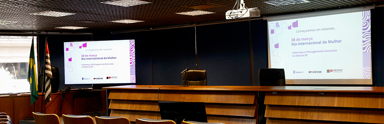 A imagem é uma foto panorâmica, interna, de um auditório. Há duas telas de projeção, uma no canto esquerdo e a outra no direito, ambas com o título: 08 de março: Dia internacional da Mulher. Ao lado da tela, à esquerda, há duas bandeiras, sendo uma do Brasil e a outra do estado de São Paulo. Já em frente às telas, estão a arquibancada e os assentos da plateia. Ao fundo, a parede é azul-escura. 