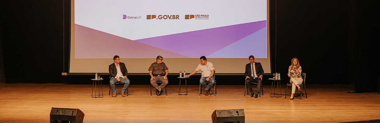 Na imagem, há um palco com cinco pessoas sentadas em cadeiras individuais, posicionadas em linha reta e voltadas para a plateia. Elas estão diante de um grande telão que exibe logotipos, incluindo o do Detran-SP e o do GOV.BR, além do nome 