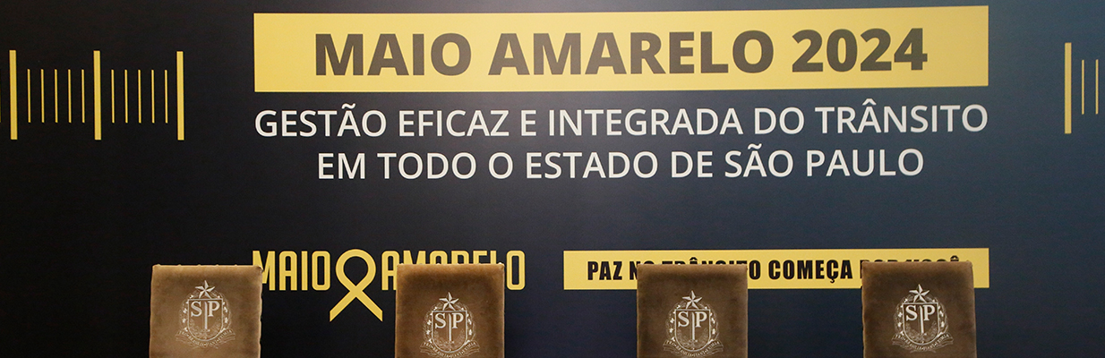 A imagem é uma foto com quatro cadeiras de veludo marrom à frente de um painel. Em cada uma das cadeiras há um bordado do brasão do estado de São Paulo. Atrás das cadeiras, no painel, está escrito: Maio Amarelo 2024: gestão eficaz e integrada do trânsito em todo o estado de São Paulo. 