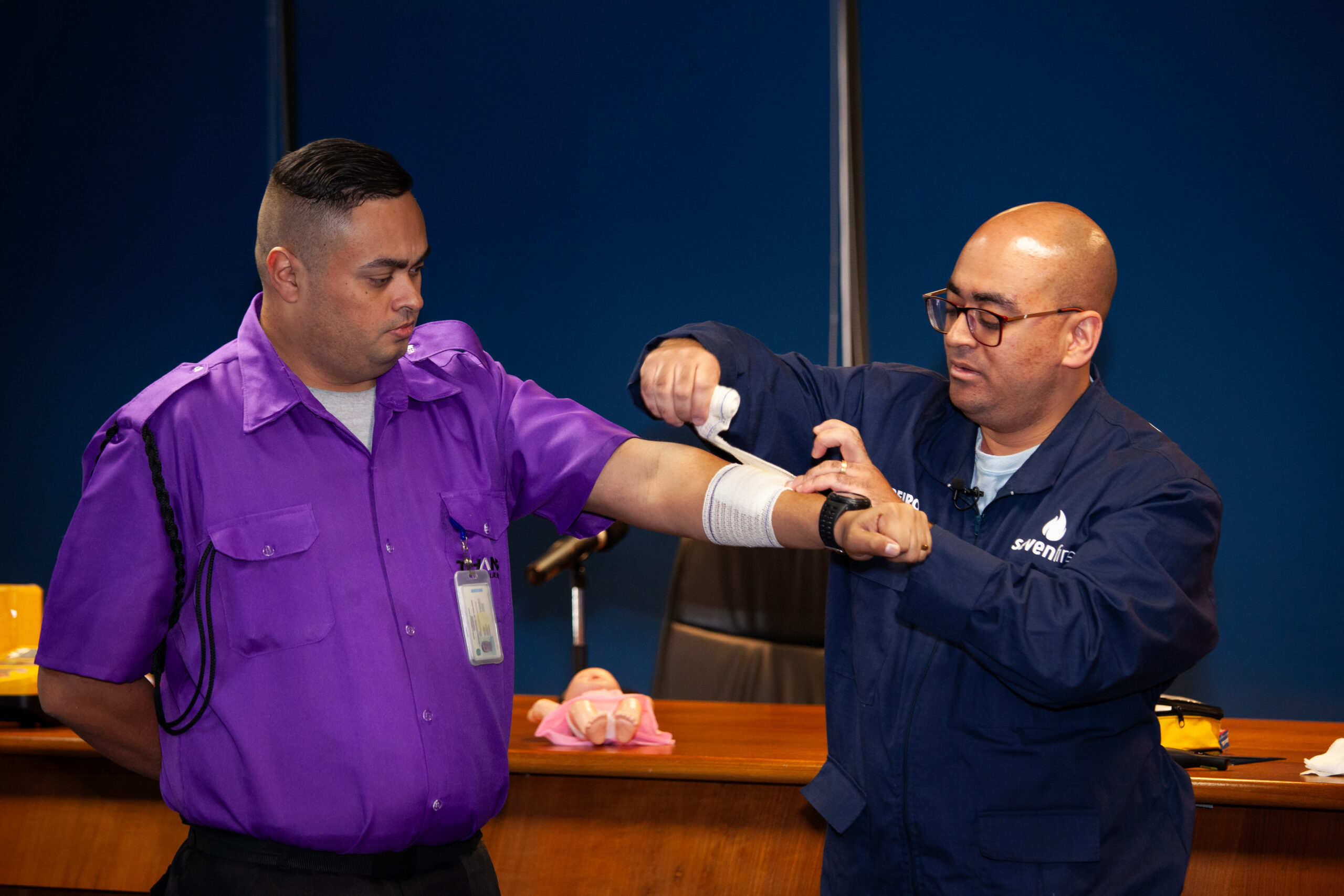 A imagem é uma foto. Em frente à bancada de um auditório, dois homens performam a ação de fazer um curativo: um deles estende o braço esquerdo, enquanto o outro enfaixa uma parte de seu antebraço.