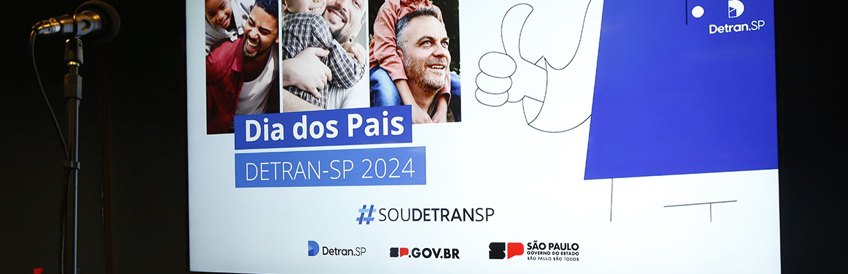 A imagem é uma foto. Em um ambiente escuro, há um telão exibindo um slide com o título “Dia dos Pais, Detran-SP 2024”. Em frente ao telão, há um microfone apoiado por um suporte vertical.