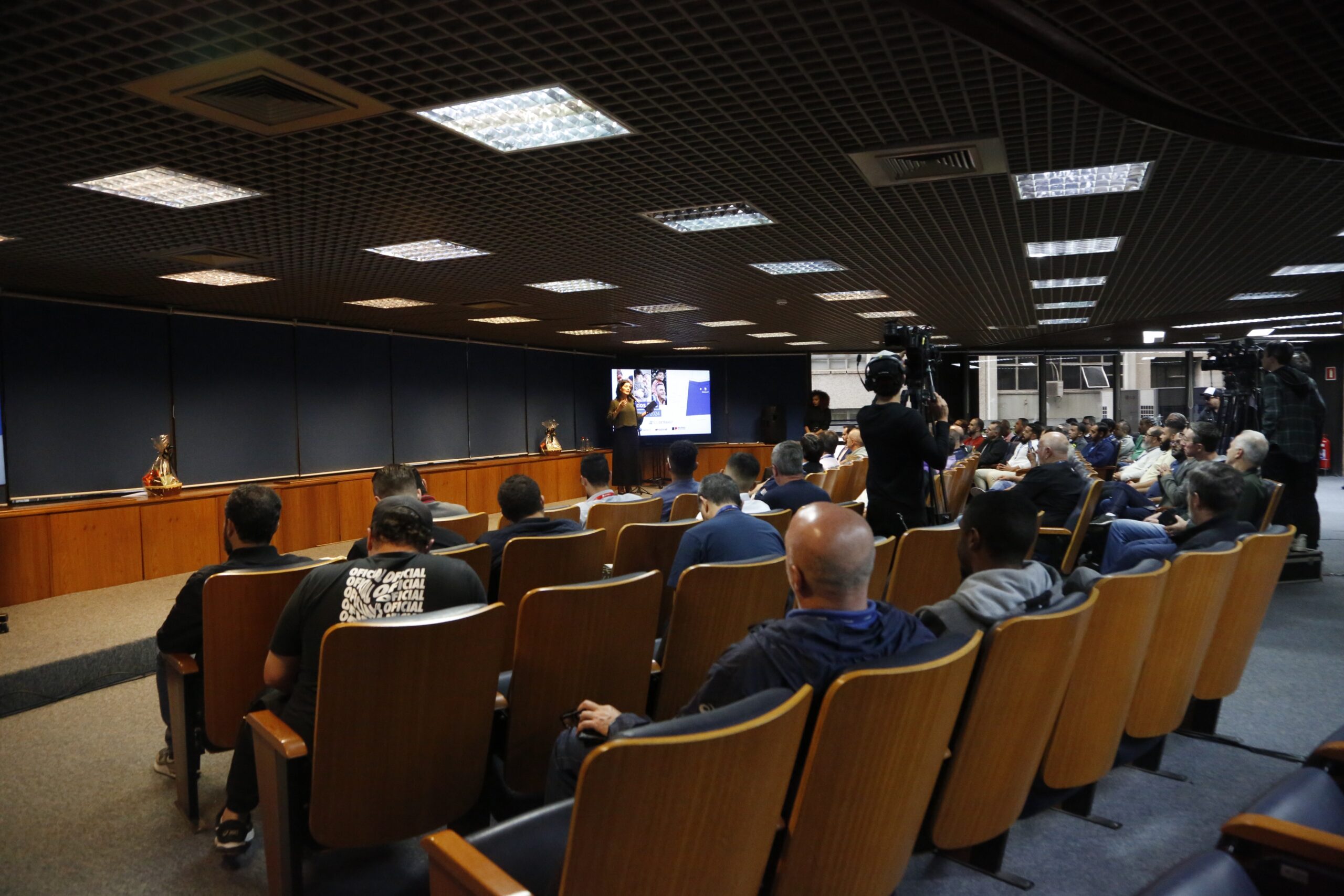 A imagem é uma foto tirada do fundo de um
								auditório. Há uma
								plateia composta por homens sentados em cadeiras de madeira. Na frente do auditório, uma mulher fala no microfone ao lado de um telão,
								