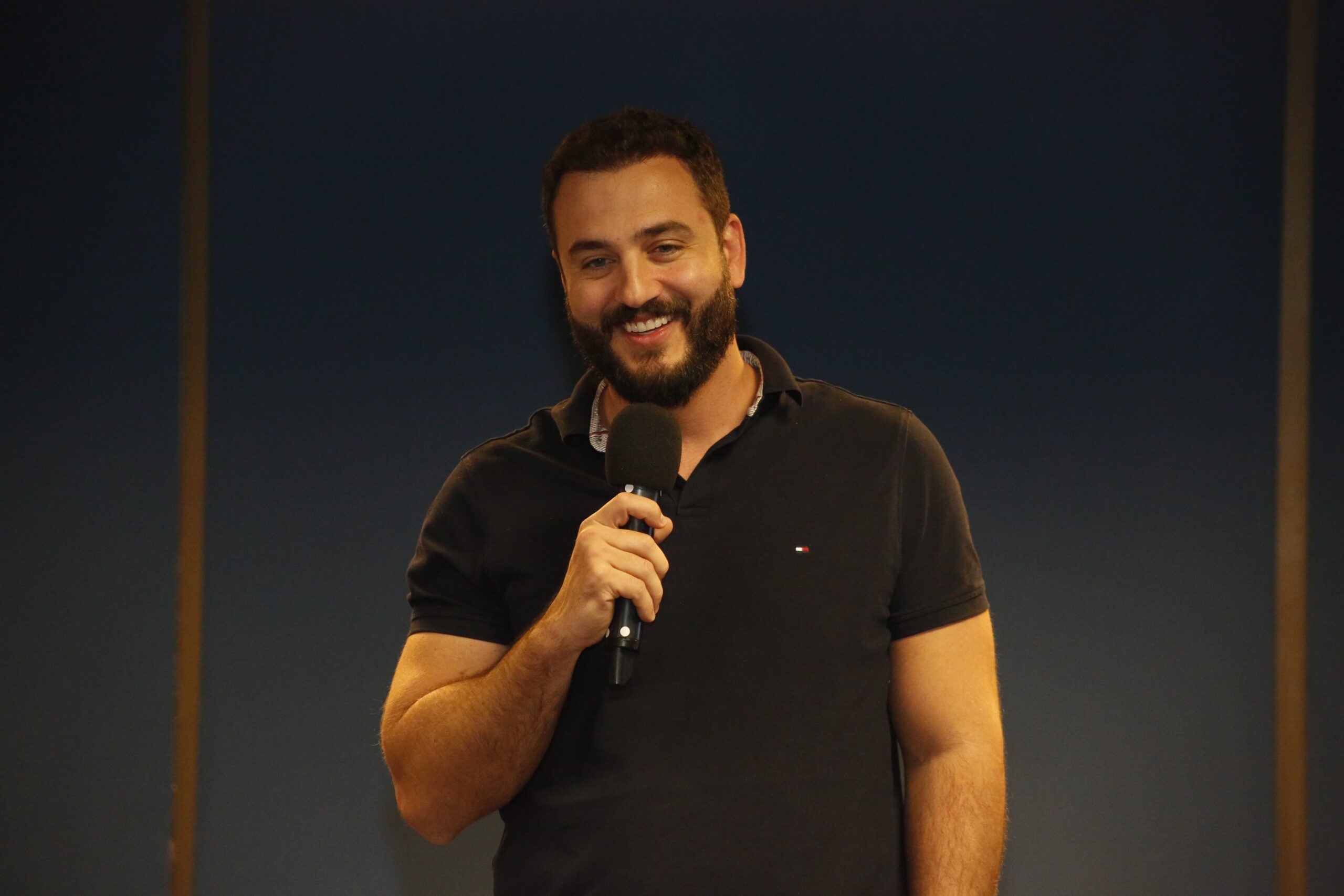 A imagem é uma foto de Eduardo Aggio, diretor-presidente do Detran-SP. Ele está sorrindo, segurando um microfone com uma das mãos. 