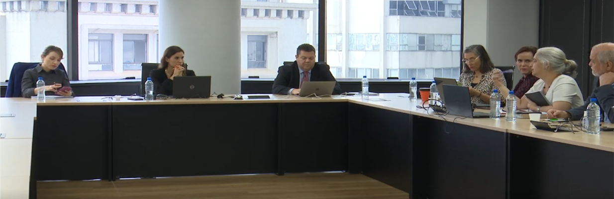 A imagem é uma foto. Em uma sala de auditório, várias pessoas com vestimentas formais estão sentadas em volta de uma bancada retangular. Sobre a bancada, há notebooks e garrafas d’água.