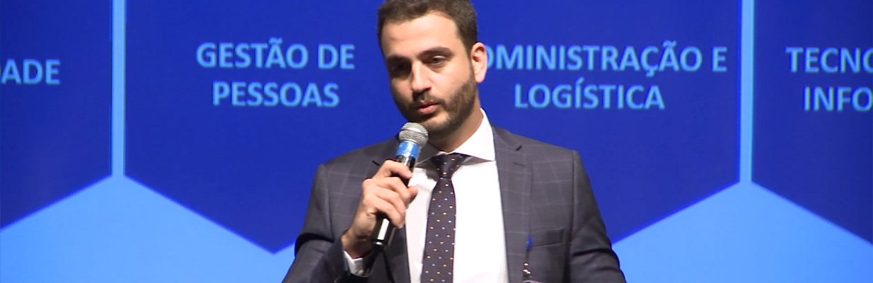 Na imagem, o diretor-presidente do Detran-SP, Eduardo Aggio, aparece falando com um microfone. Ele veste um terno escuro com uma camisa branca e uma gravata estampada. Sua expressão é séria e concentrada enquanto fala. Ao fundo, há um painel azul dividido em três seções, cada uma com uma legenda em letras maiúsculas brancas: “QUALIDADE”, “GESTÃO DE PESSOAS”, “ADMINISTRAÇÃO E LOGÍSTICA”, e “TECNOLOGIA DA INFORMAÇÃO”. Cada título está dentro de uma forma hexagonal que compõe um padrão maior de hexágonos que se conectam. Não é possível ver os detalhes ou o conteúdo de cada seção do painel, pois o homem está posicionado em frente a ele.