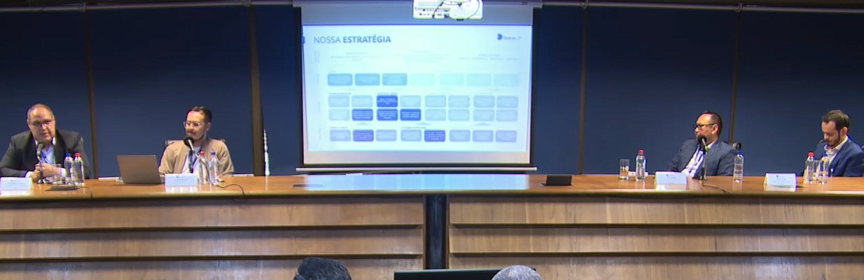 A imagem é uma foto horizontal. Da esquerda para a direita, estão atrás de uma arquibancada: Eduardo Gomes, Superintendente de São José dos Campos do Detran-SP; Silvio Tanaka Fonseca, Gerente do Produto & Líder LabInova/Detran-SP; José Iberê Fernandes Junior, da Assessoria de Governança e Inteligência Corporativa; e, por fim, Lucas Papais, da Diretoria de Atendimentos. No centro da imagem há uma tela de projeção, com um slide de conteúdo que varia em tons de azul e cinza.