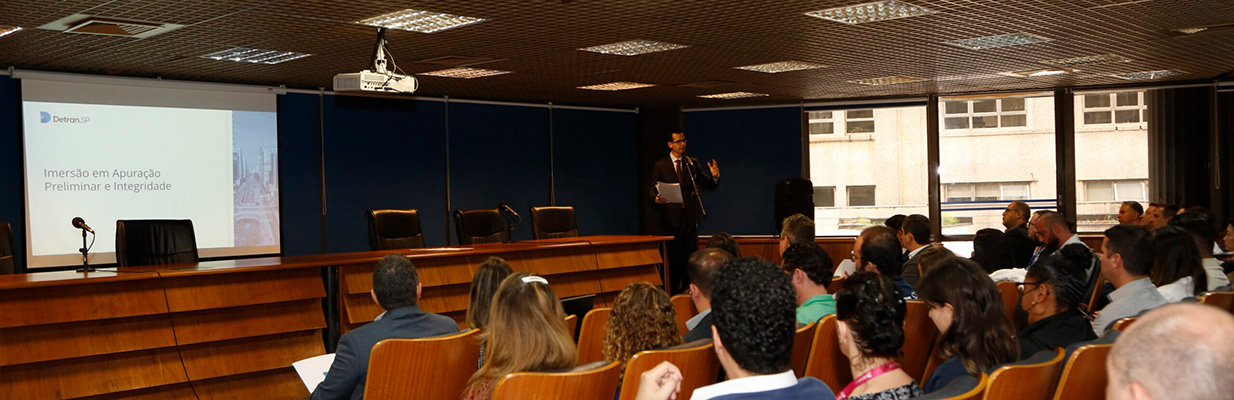 A imagem mostra um auditório. À direita, em frente a um grupo de pessoas sentadas, há um homem de pé fazendo uma apresentação. Ele veste um terno escuro com uma camisa branca e gesticula com a mão direita. Atrás dele, há uma tela de projeção grande que exibe uma apresentação em PowerPoint. A tela tem um cabeçalho que diz “Detran-SP”, seguido pelo subtítulo “Imersão em Apuração Preliminar e Integridade”. O fundo da tela de projeção mostra uma imagem de uma estrutura arquitetônica. O público está sentado em fileiras de cadeiras estofadas na cor creme, ouvindo atentamente. A sala tem paredes azuis-escuras e teto com telhas de absorção acústica. Ao fundo, há janelas que mostram a lateral de um prédio ao lado e o céu claro.